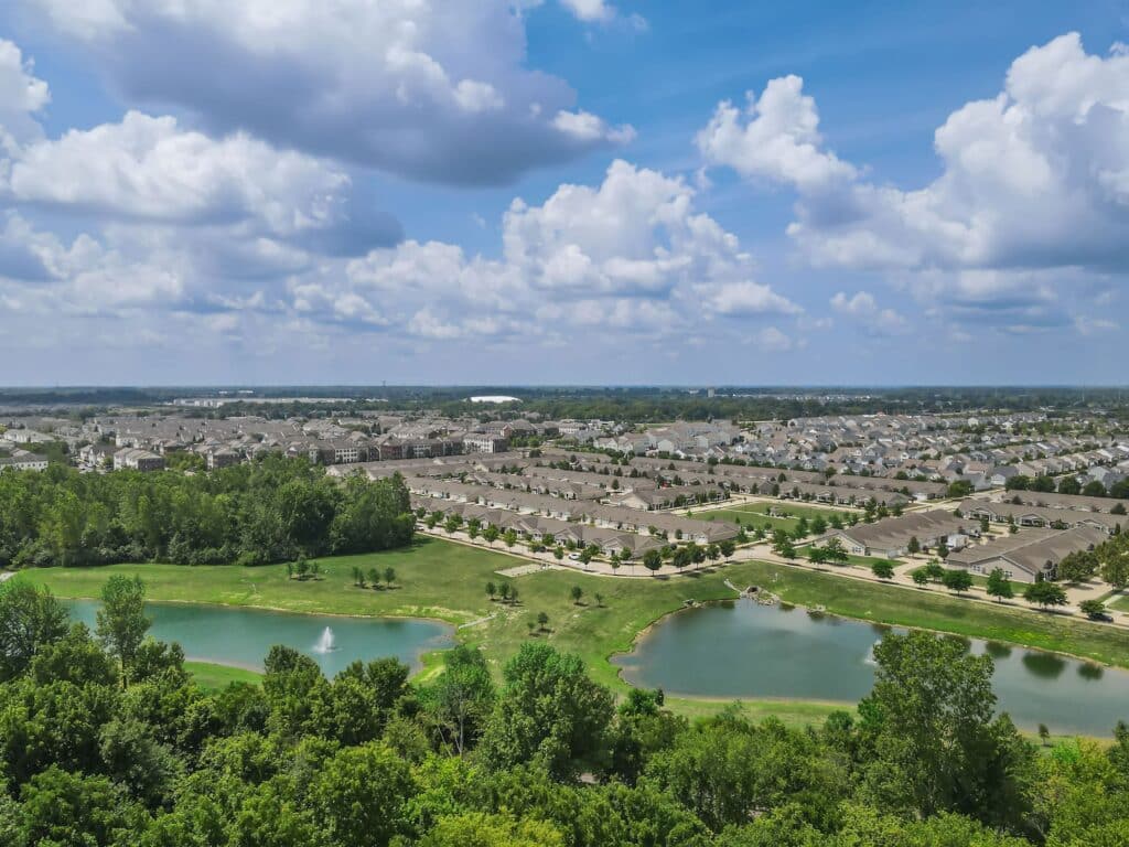 Aerial view of a suburban neighborhood with rows of houses, green trees, two small ponds, and a fountain under a partly cloudy sky.