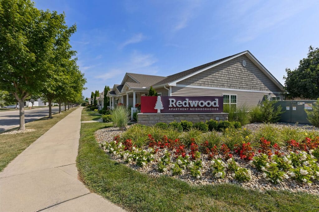 A landscaped sidewalk leads to a single-story building with a sign reading "Redwood Apartment Neighborhoods" in front, surrounded by flowers and greenery.