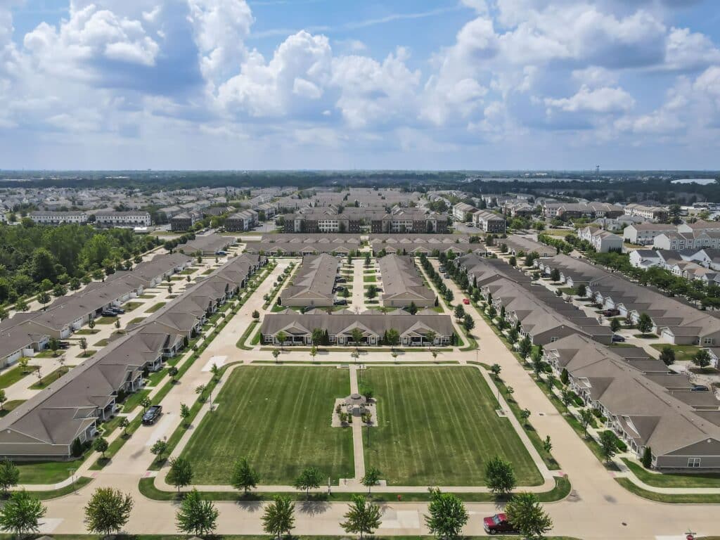 Aerial view of a suburban neighborhood with rows of identical houses, green lawns, and a central park area under a partly cloudy sky.