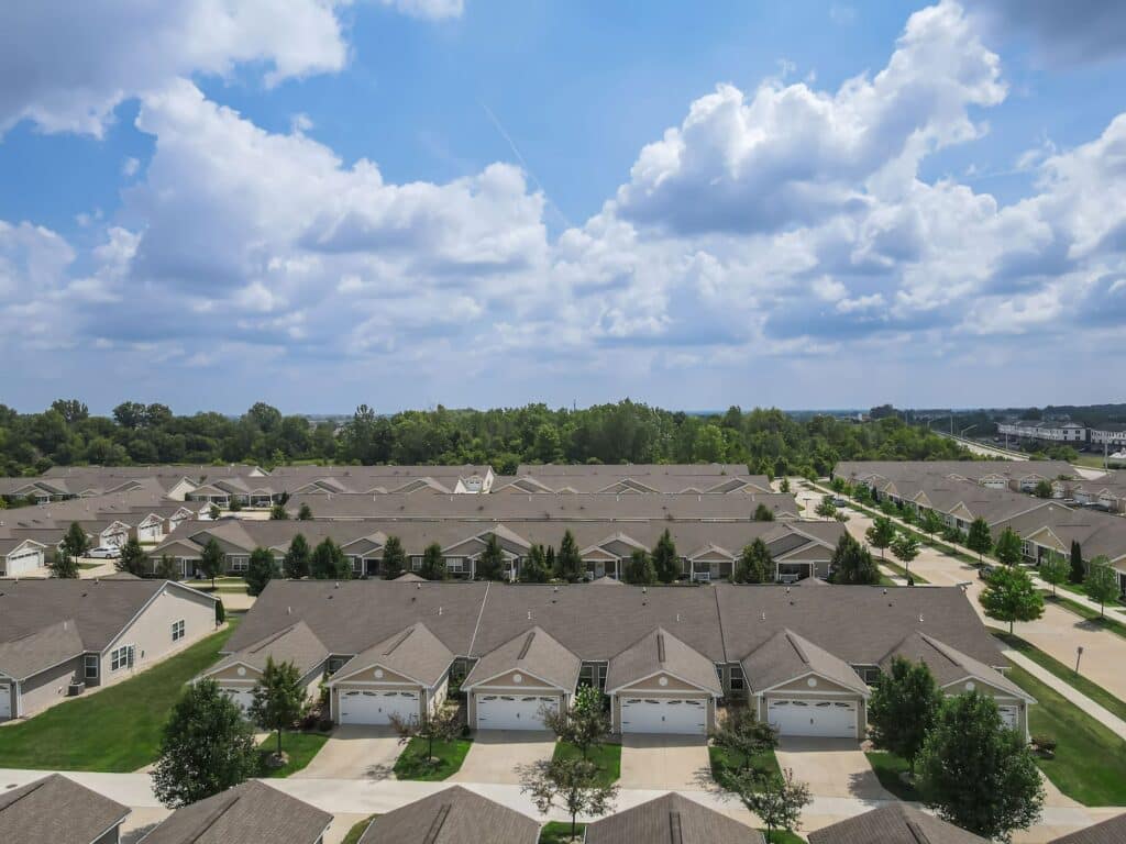 Aerial view of a suburban neighborhood showing rows of single-story houses with garages, green lawns, and tree-lined streets under a partly cloudy sky.