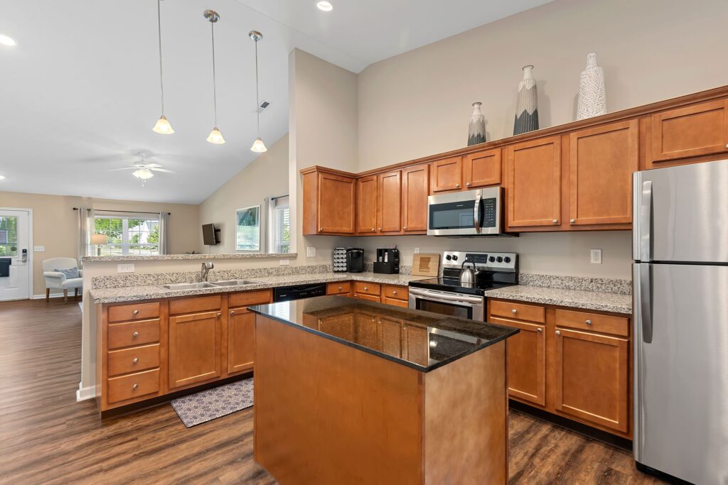 Modern kitchen with wooden cabinets, granite countertops, stainless steel appliances, and a central island; open layout leads to a living area with large windows.