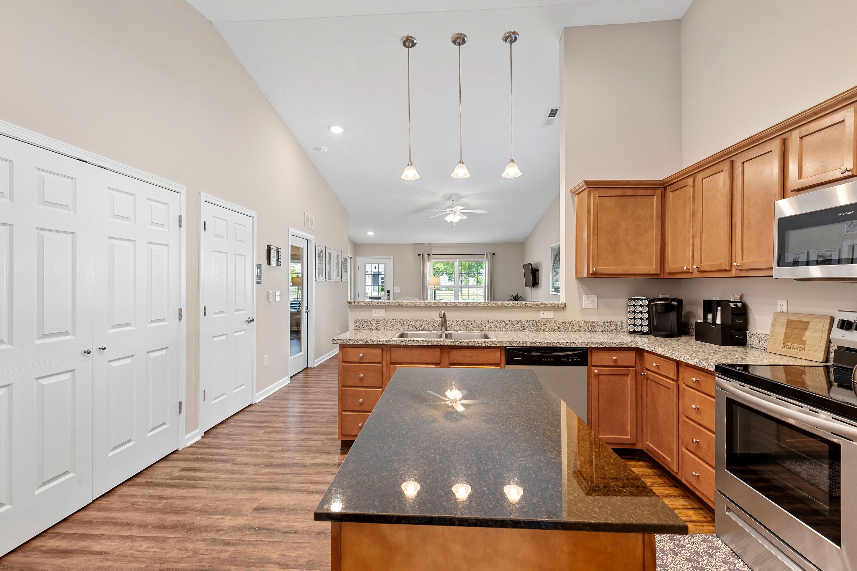Modern kitchen with wood cabinets, granite countertops, center island, stainless steel appliances, pendant lights, and a view into the living area with ceiling fan and large windows.
