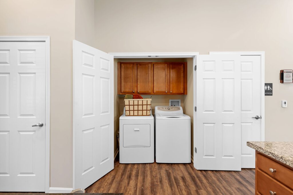 A laundry area with a white washer and dryer, wooden cabinets above, and double doors open to reveal the appliances; wood floor and beige walls.