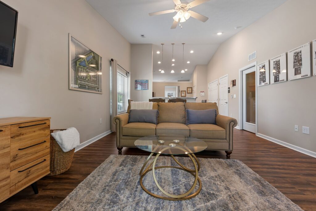 A modern living room with a tan sofa, glass coffee table, blue rug, wooden dresser, wall art, and ceiling fan, featuring hardwood floors and neutral-colored walls.