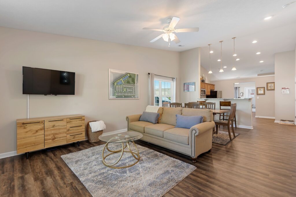 A modern open-concept living and dining area with a beige sofa, glass coffee table, TV on the wall, wooden furniture, and a view of the kitchen in the background.