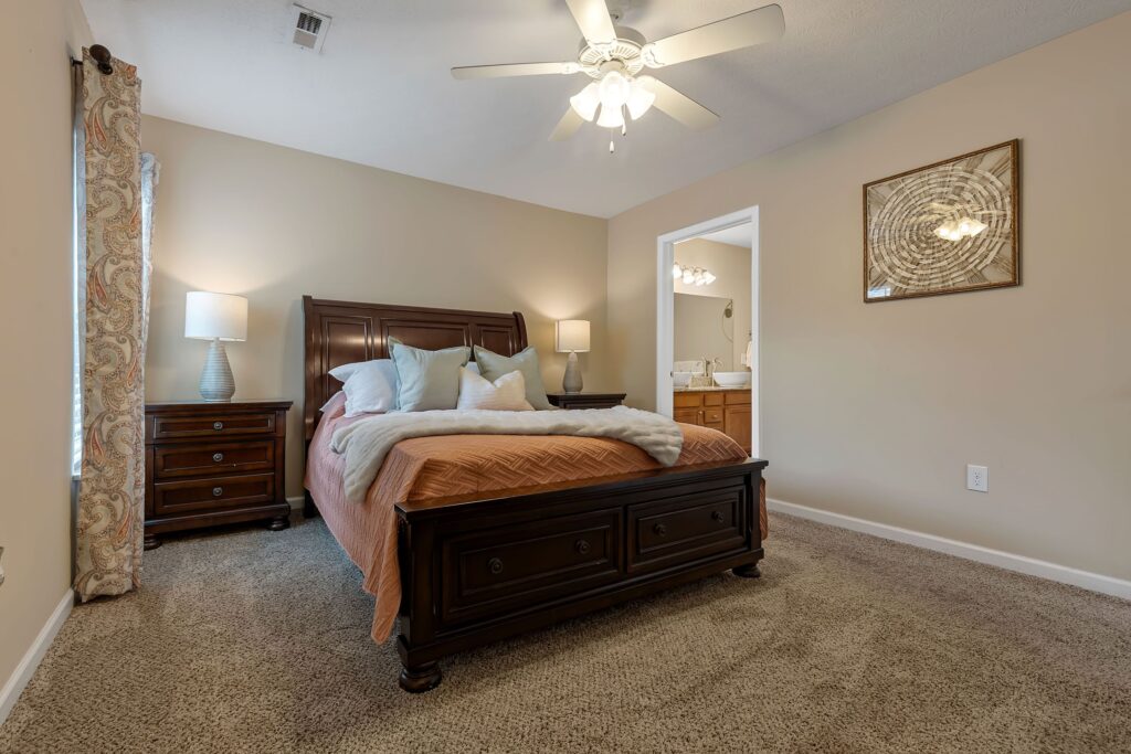 A neatly made bed with a dark wooden frame sits in a carpeted bedroom with beige walls, two nightstands, and a ceiling fan; an open door leads to a bathroom.