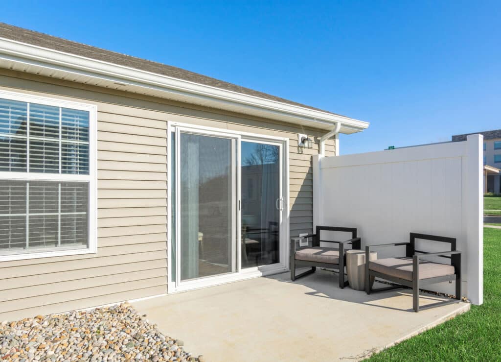 A small patio with two cushioned chairs, a white privacy screen, sliding glass doors, and beige siding on the house, under a clear blue sky.