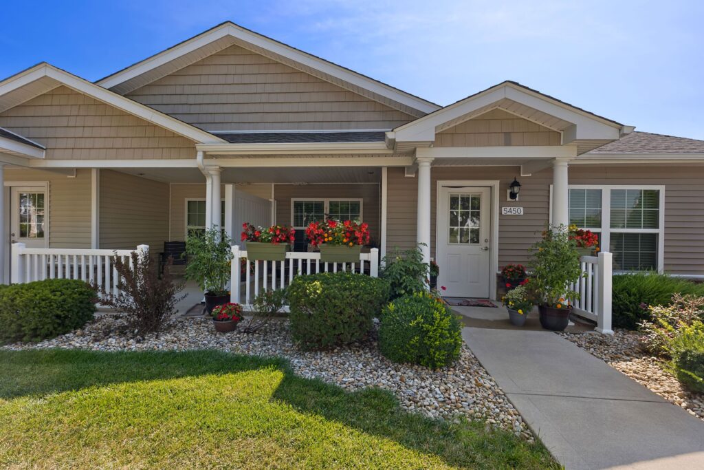 Single-story house with beige siding, a covered front porch, white railings, flower boxes, potted plants, and a stone-lined walkway surrounded by green shrubbery and lawn.