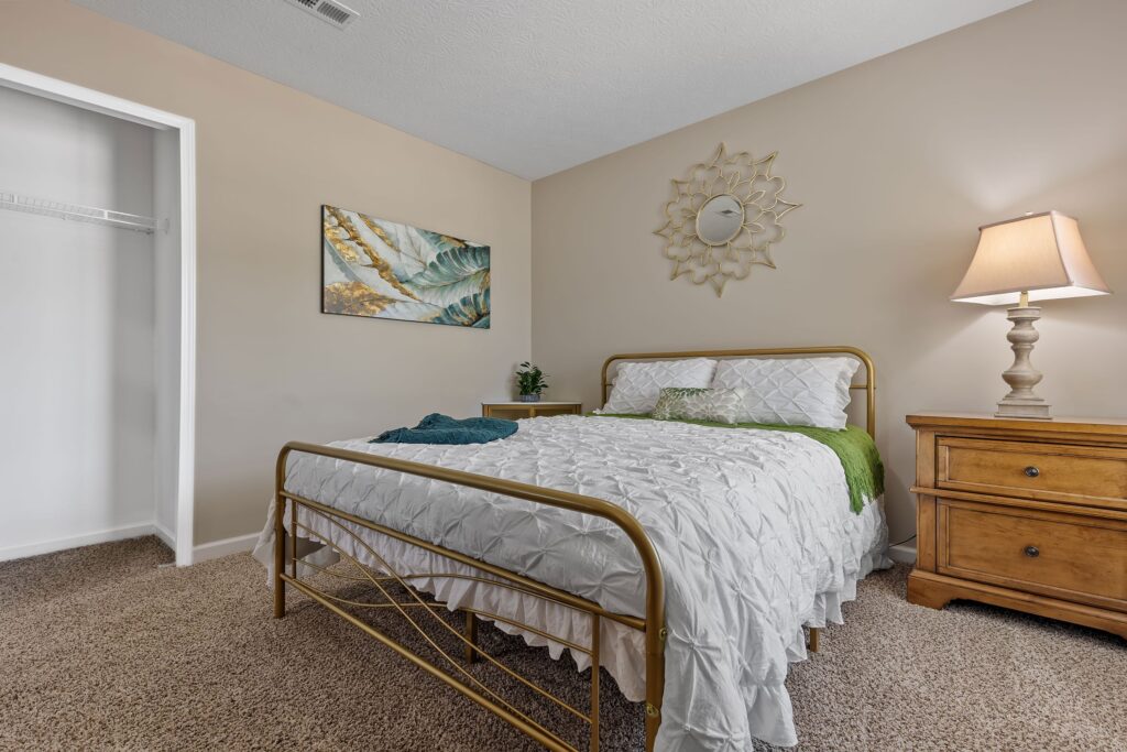 A neatly made bed with a white comforter and gold frame sits beside a wooden nightstand with a lamp in a carpeted bedroom with beige walls and an open closet.