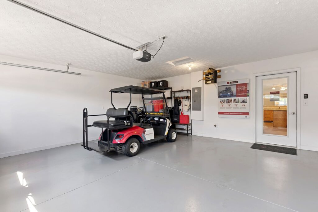 A red and white golf cart is parked in a clean, spacious garage with white walls, organized shelves, wall posters, and a door leading to another room.