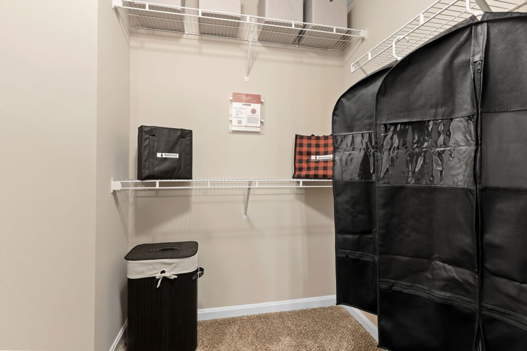 A walk-in closet with wire shelves, storage bins, a black laundry hamper, carpeted floor, and a black portable wardrobe.