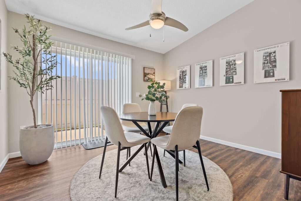 A modern dining area with a round table, four chairs, a potted tree, framed floor plans on the wall, and vertical blinds letting in natural light.