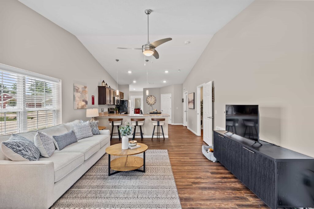Modern open-concept living room and kitchen with neutral tones, hardwood floors, a sofa, coffee table, barstools, and a TV console. Large windows provide natural light.