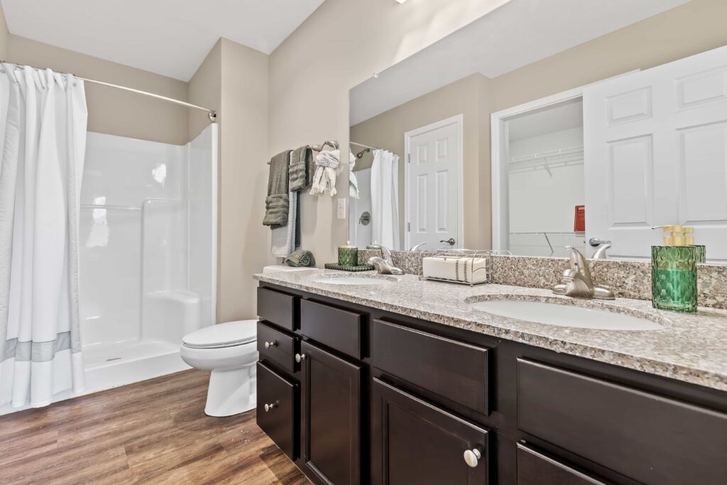 Modern bathroom with dark wood cabinets, granite countertop, dual sinks, a toilet, and a walk-in shower with a white curtain. Towels and toiletries are neatly arranged.