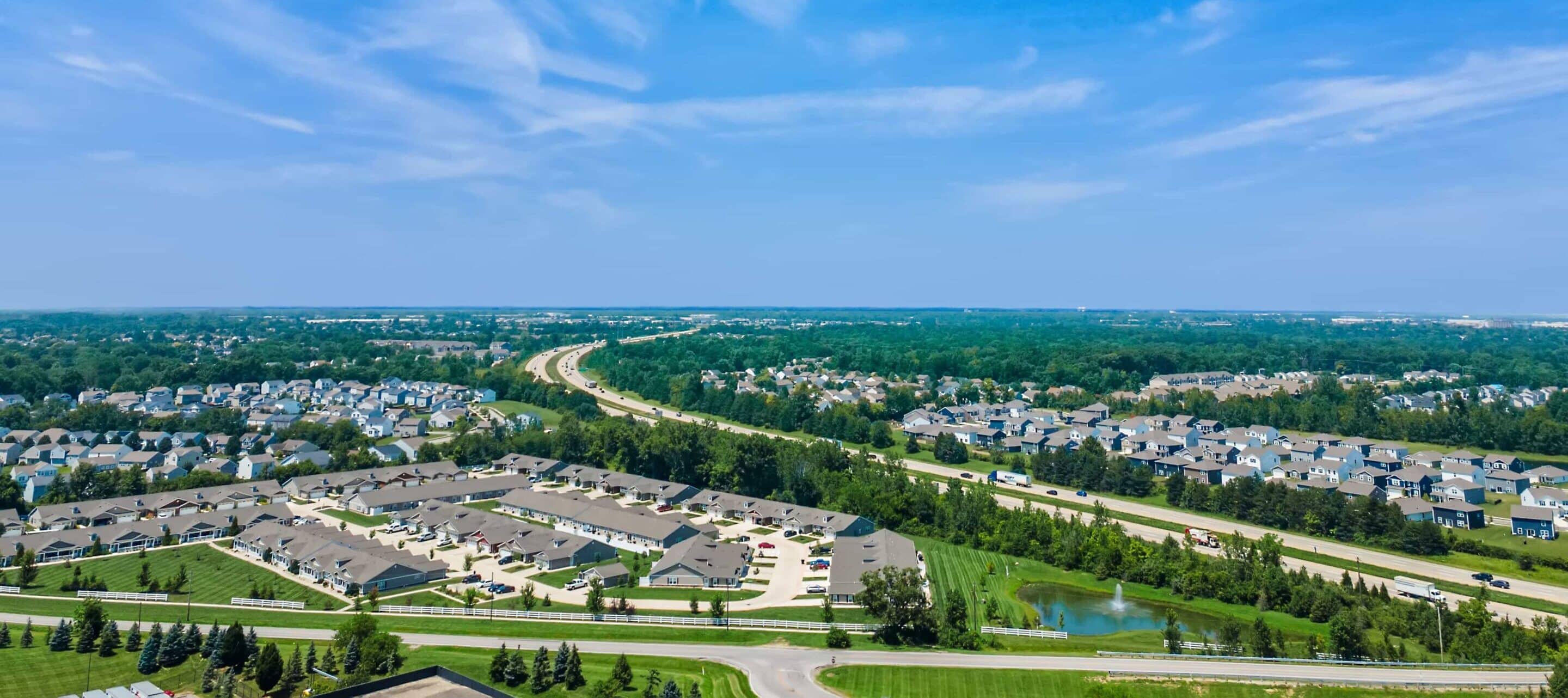 Aerial view of a suburban neighborhood with rows of houses, roads, parking lots, and green spaces under a blue sky with scattered clouds.