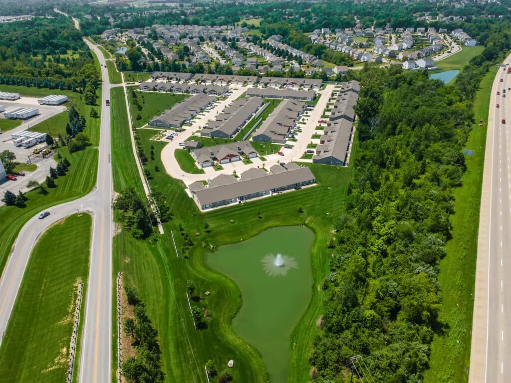 Aerial view of a suburban neighborhood with rows of houses, a pond with a fountain, surrounding greenery, and nearby roads including a highway.