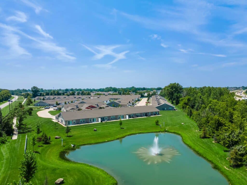 Aerial view of a residential neighborhood with single-story buildings, a pond with a fountain, green lawns, and surrounding trees under a blue sky.