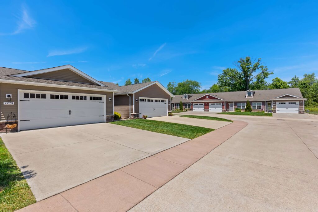 Wide concrete driveway leads to several single-story, suburban houses with attached garages under a clear blue sky.