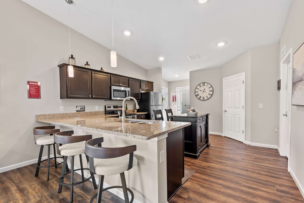 Modern kitchen with dark cabinets, granite countertops, a breakfast bar with four stools, stainless steel appliances, wood flooring, and recessed lighting.
