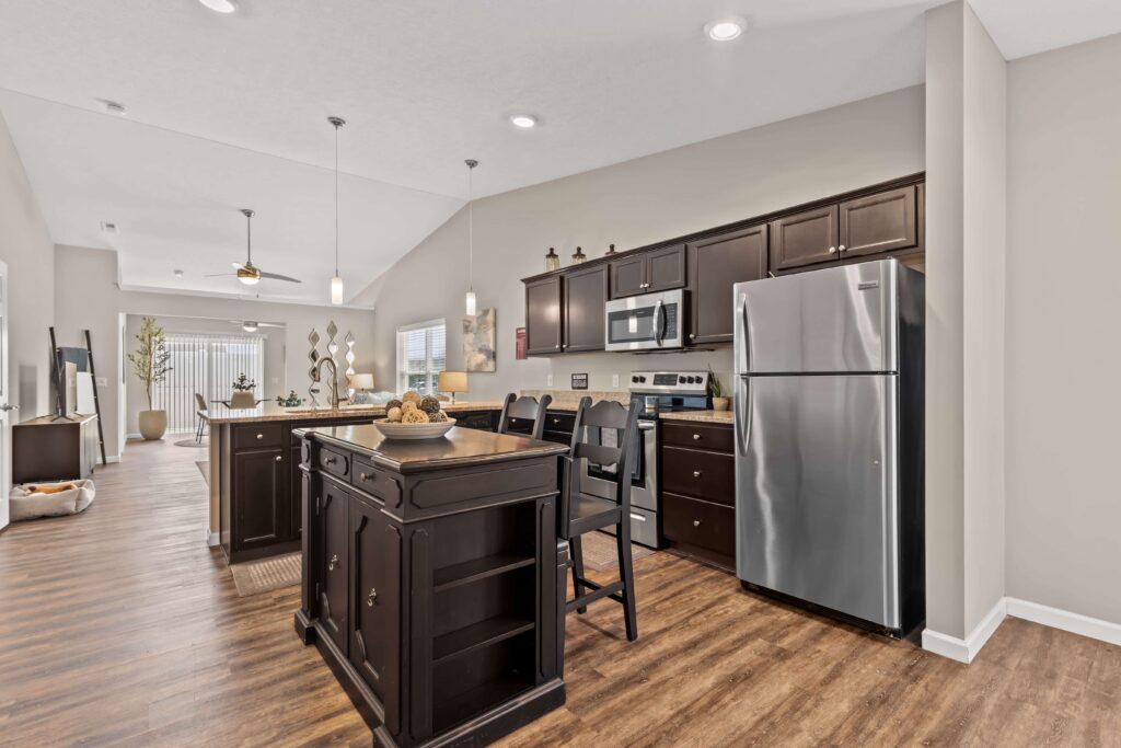 Modern kitchen with dark cabinets, stainless steel appliances, a central island with seating, and wood-style flooring, open to a living and dining area.