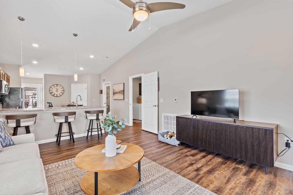 Modern living room with wood flooring, a sofa, coffee table, TV on a dark stand, and kitchen with bar stools in the background. Ceiling fan and neutral decor are visible.