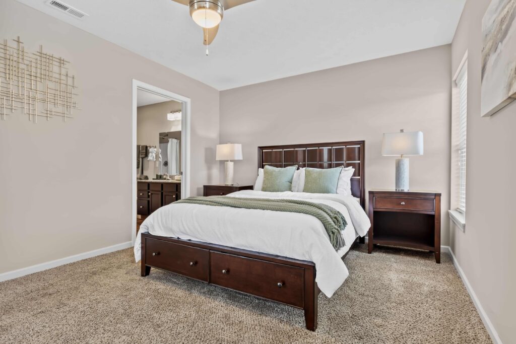 A neatly made bed with white bedding and green pillows in a carpeted bedroom with two nightstands and lamps; a bathroom is visible through an open door.