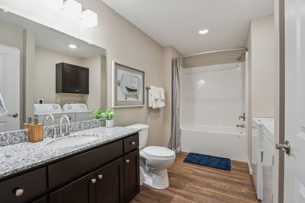 Modern bathroom with dark wood vanity, granite countertop, a toilet, washer and dryer, bathtub with shower curtain, wall art, and wood-style flooring.