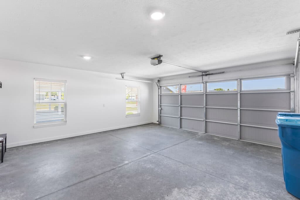 Clean, empty two-car garage with concrete floor, white walls, two windows, ceiling lights, and a large garage door with windows. A blue bin is placed on the right side.