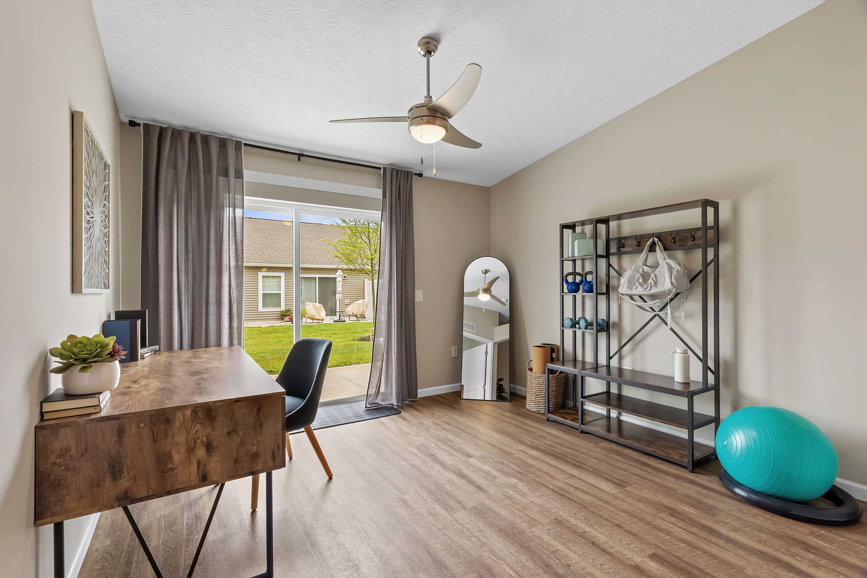 A home office with a wooden desk, black chair, shelving unit, large mirror, exercise ball, and sliding glass doors leading to a backyard area.