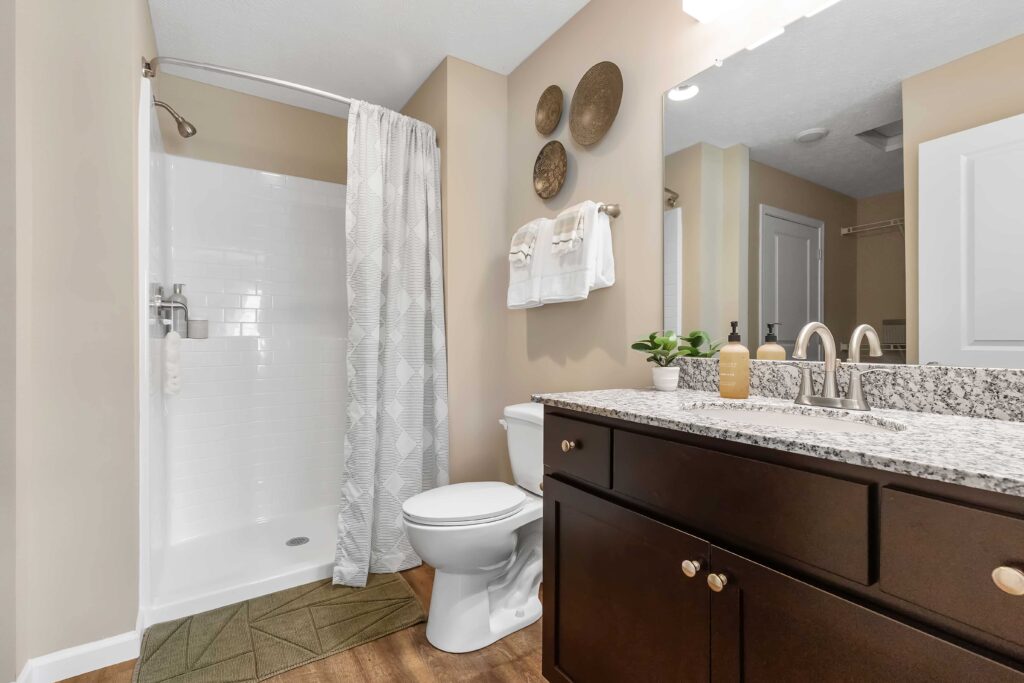 Modern bathroom with a glass-less shower, beige walls, granite countertop, dark wood vanity, and decorative wall accents above the toilet.