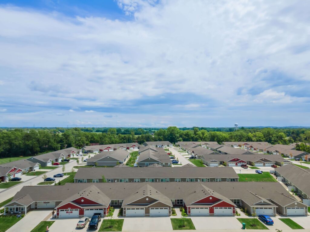 Aerial view of a suburban neighborhood with rows of single-story houses, driveways, grassy areas, and parked cars under a partly cloudy sky.