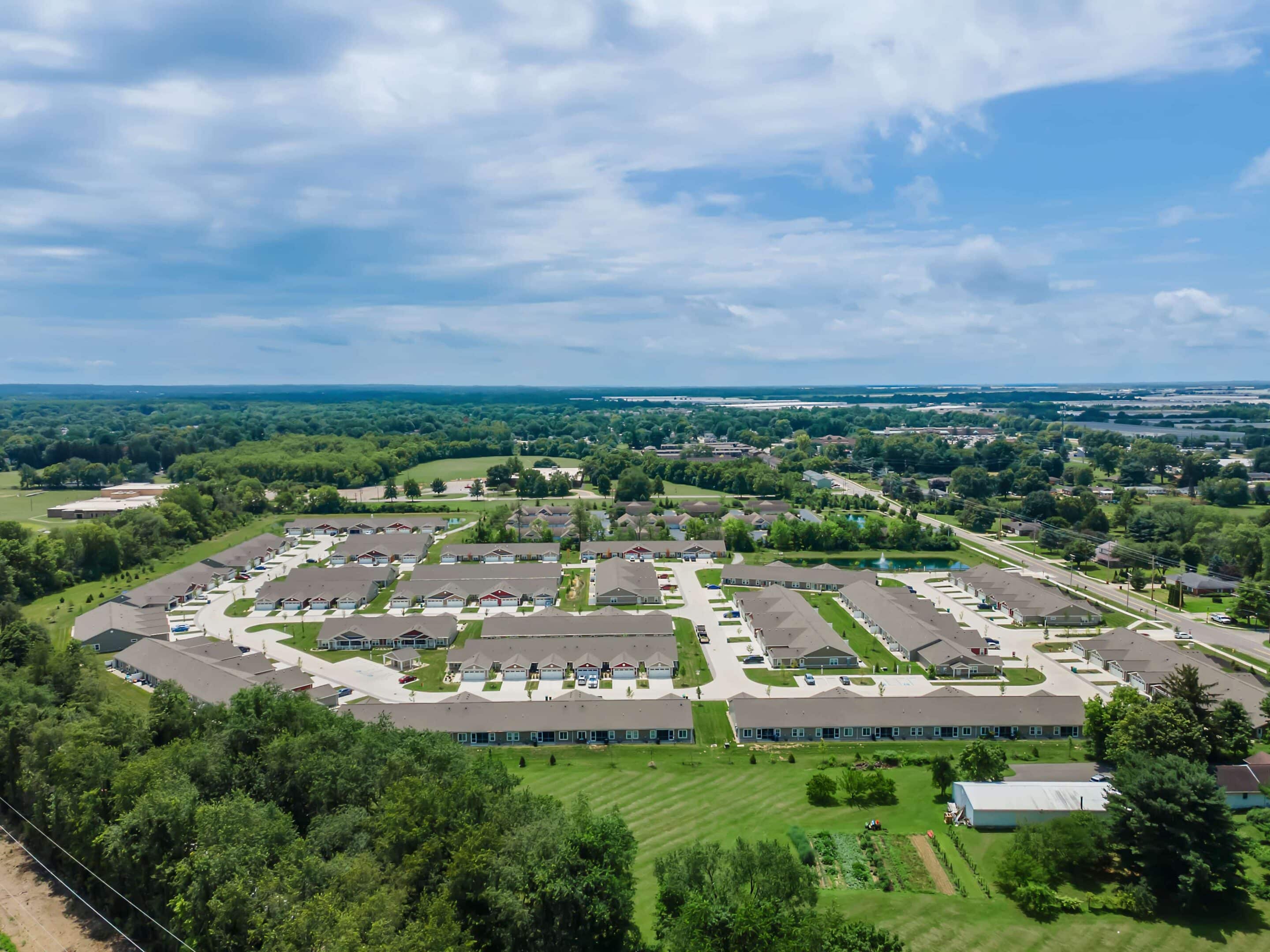 Aerial view of a suburban residential neighborhood with rows of houses, green lawns, and surrounding trees under a partly cloudy sky.