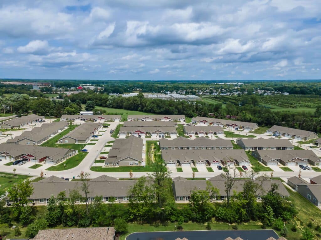 Aerial view of a suburban residential neighborhood with rows of houses, streets, and green spaces under a partly cloudy sky.