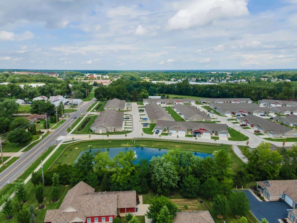 Aerial view of a suburban neighborhood with rows of houses, a small pond, green trees, and a road running alongside the area under a partly cloudy sky.