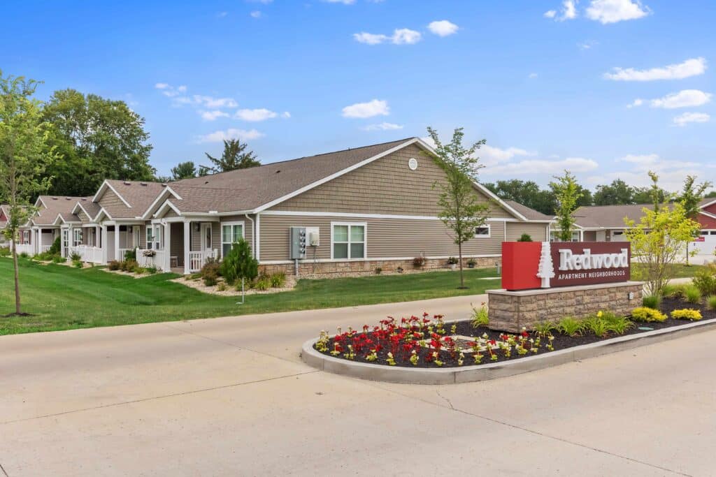 Single-story apartment buildings with manicured lawns and a red "Redwood Apartment Neighborhoods" sign surrounded by flowers at the entrance.