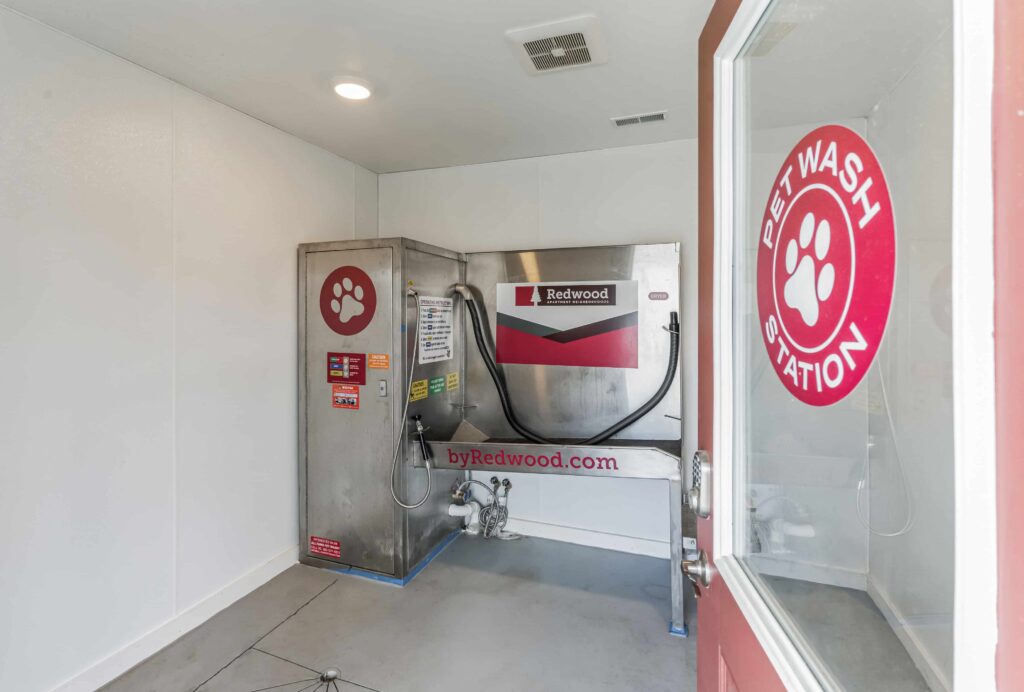 A stainless steel pet wash station with hoses and a red paw print logo in a clean, white room viewed through an open door.