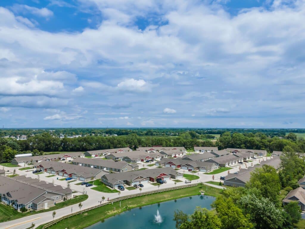 Aerial view of a suburban neighborhood with rows of houses, green lawns, and a small pond with a fountain under a partly cloudy sky.