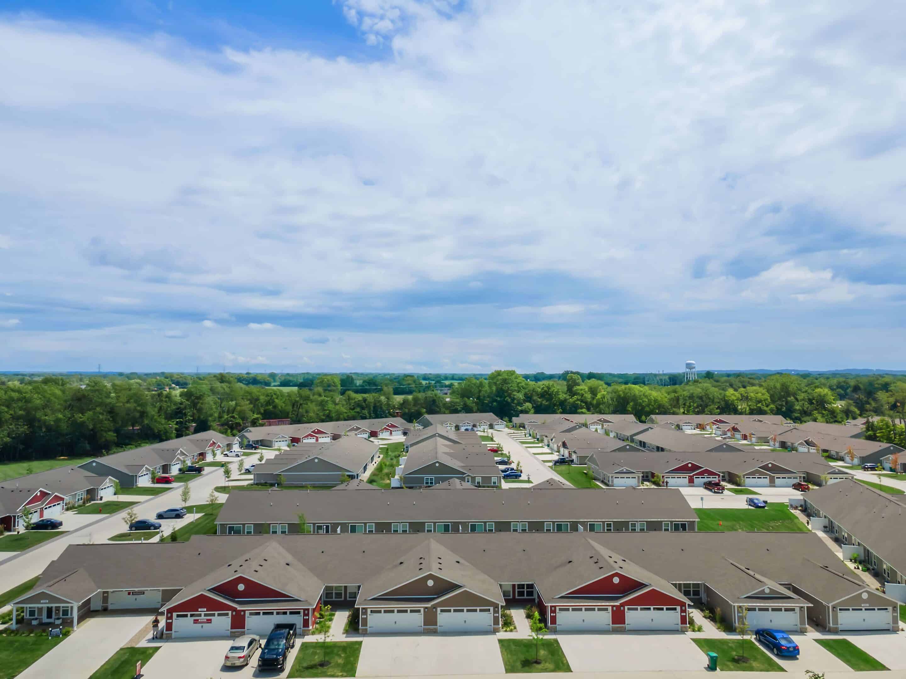 Aerial view of a suburban neighborhood with rows of single-story houses, driveways, grassy areas, and parked cars under a partly cloudy sky.