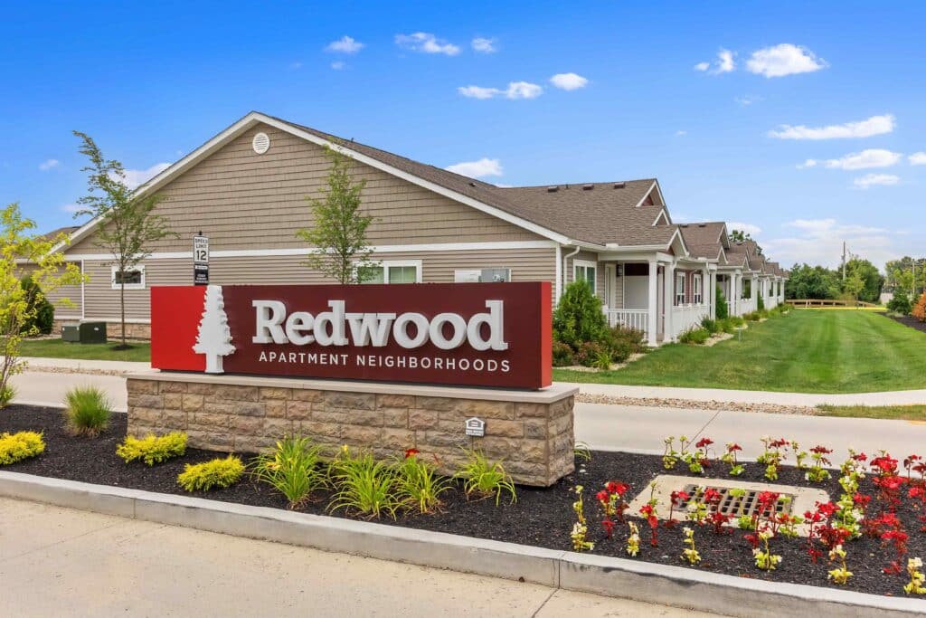 A sign reading "Redwood Apartment Neighborhoods" stands in front of single-story apartment buildings with landscaped greenery under a blue sky.