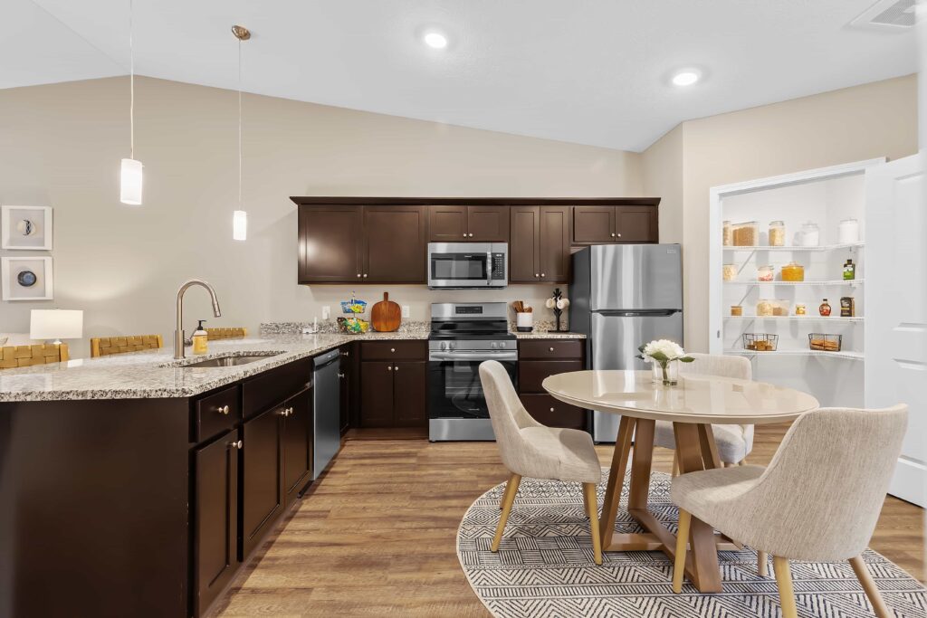 Modern kitchen with dark cabinets, stainless steel appliances, a round dining table with four chairs, and an open pantry stocked with jars and baskets.