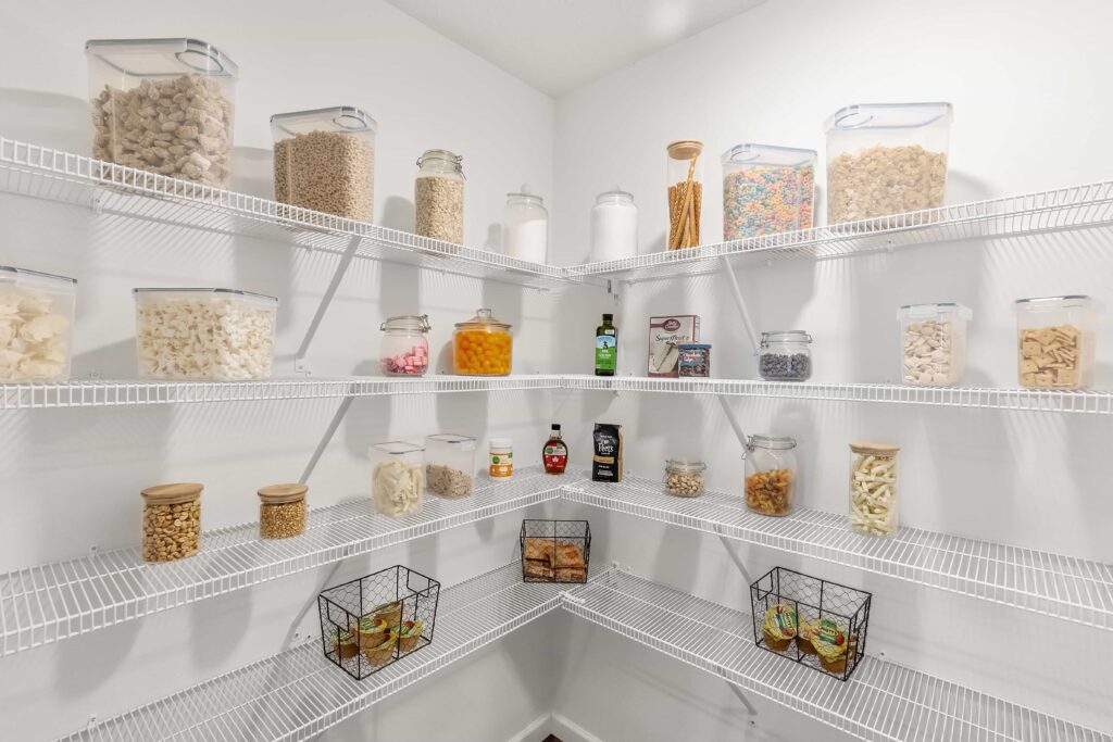 A neatly organized pantry with clear containers holding cereals, pasta, snacks, and baking supplies on white wire shelves against white walls.