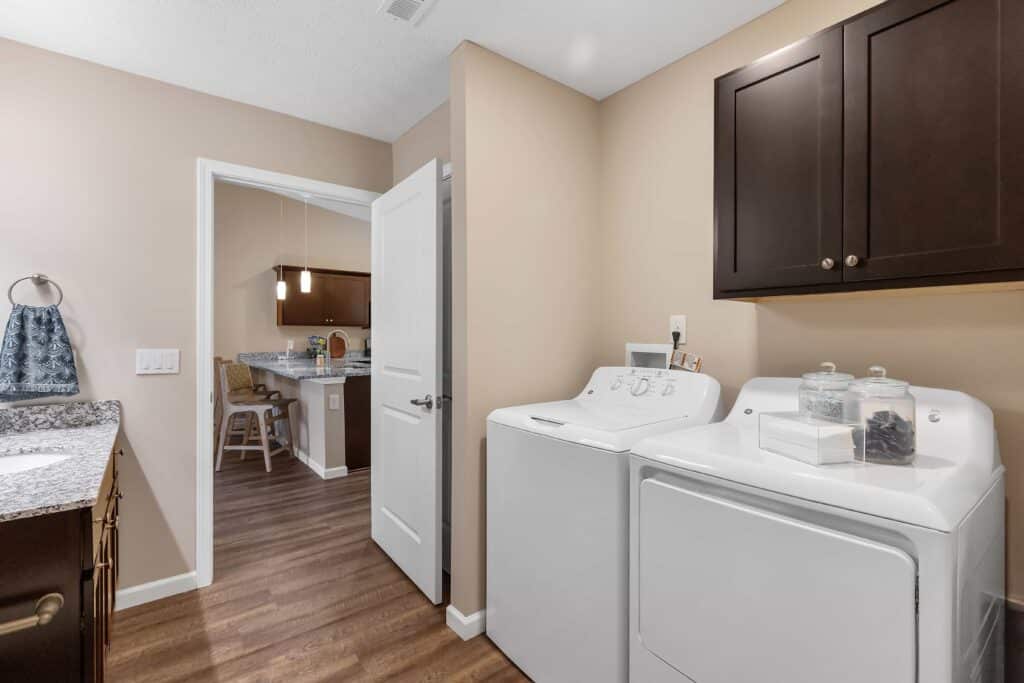 A laundry area with a washer and dryer, brown cabinets, glass jars on top, and a view into a kitchen and barstools in the background.