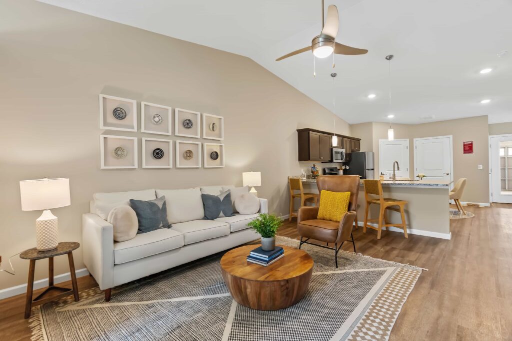 Modern living room and kitchen with neutral tones, a white sofa, round wooden coffee table, decorative wall art, ceiling fan, and wooden dining set on hardwood floors.