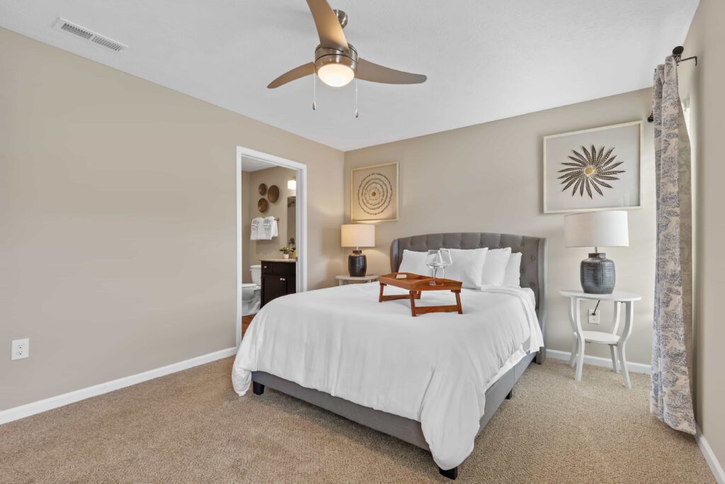 Bedroom with beige walls, a ceiling fan, a bed with white bedding, two nightstands with lamps, decorative wall art, and a tray on the bed; bathroom visible through open door.