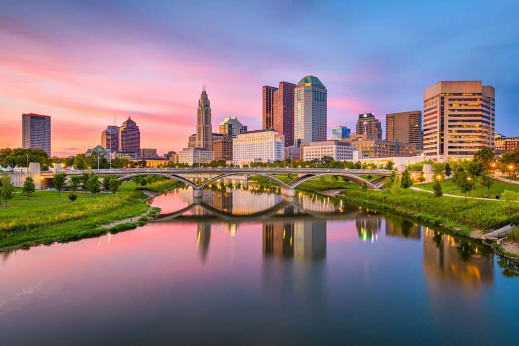 City skyline with modern high-rise buildings, a bridge, and a river in the foreground at sunset with a colorful sky.