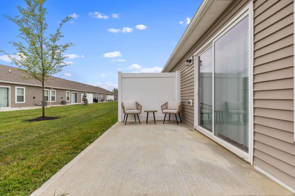 A small concrete patio with two chairs and a table, bordered by a privacy panel, outside a beige house with sliding glass doors and a grassy yard.