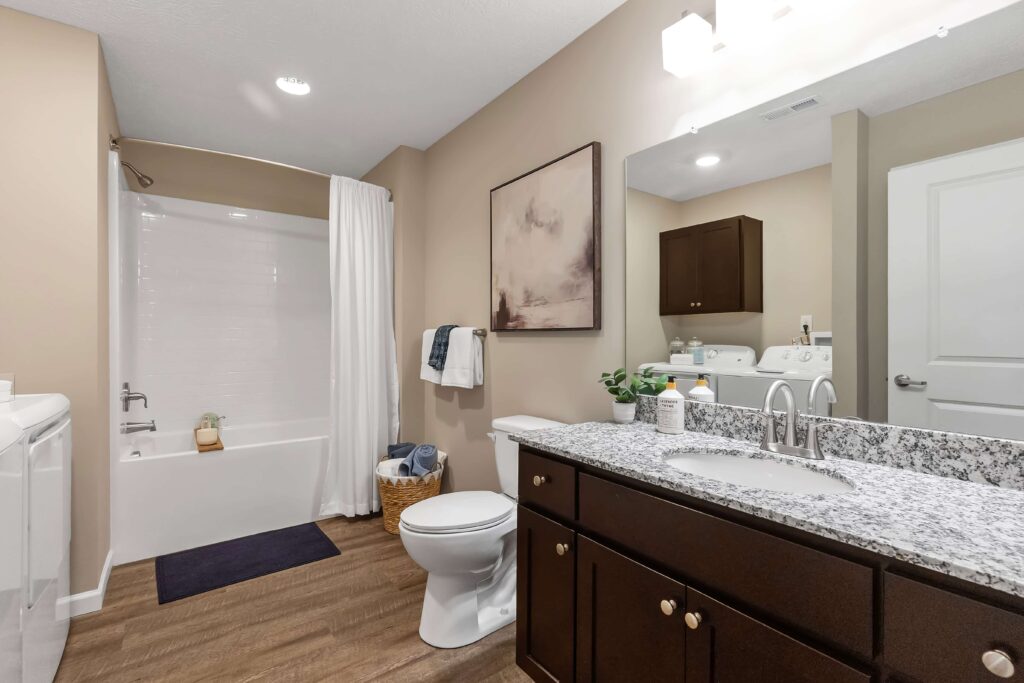 Modern bathroom with granite countertop, single sink, toilet, bathtub with white shower curtain, wooden cabinets, and laundry appliances in the background.