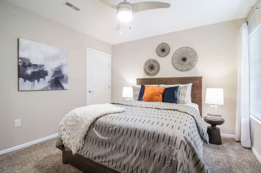 A neatly made bed with patterned bedding, two orange and blue accent pillows, bedside lamps, and abstract wall art in a bright, modern bedroom.