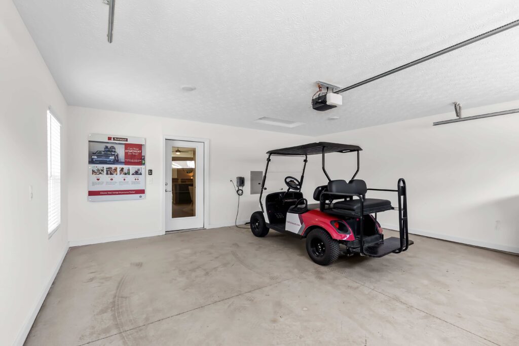 A red golf cart is parked in a clean, empty garage with white walls, two windows, and a door leading to an adjacent room.