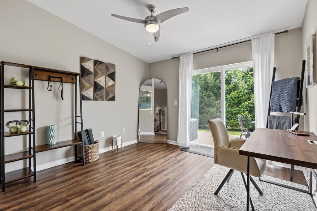 Modern home office with wooden floor, desk, chairs, tall mirror, shelving, and sliding glass doors opening to a green outdoor area.